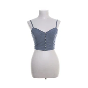 Blå croppad korsettopp med knappar - Snygg croppad korsettopp i ljusblått med smala justerbara axelband och knäppning framtill. Toppen har markerade sömmar och en liten metallogo på ena sidan. Perfekt för att skapa en cool och trendig look.