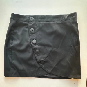 Svart omlottkjol med knappar - Supersnygg svart kort kjol från ASOS i storlek 40. Fin tulpanmodell med dekorativa (fungerande) knappar fram. Har aldrig använts, ty jag har ätit för många bullar (är för liten) och jag inte har några planer på att sluta med det. 😅  Hämtas i Stockholm eller skickas mot porto.