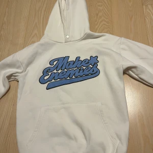 Vit hoodie blå text - Vit hoodie med stor blå broderad text på bröstet. Tröjan har huva, känguruficka och långa ärmar. Perfekt för dig som gillar streetwear och vill sticka ut med en snygg detalj framtill.