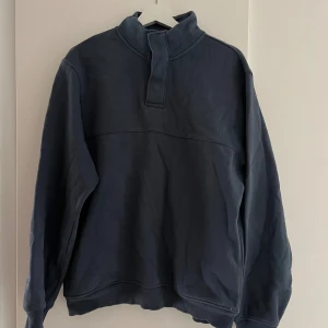 Mörkblå sweatshirt med tryckknappar NEXT - Mörkblå sweatshirt från NEXT i storlek M. Tröjan har hög krage med tryckknappar och ribbade muddar vid ärmslut och nederkant. Perfekt för lager-på-lager och chill dagar. Tillverkad i mjuk bomullsmix för extra komfort.