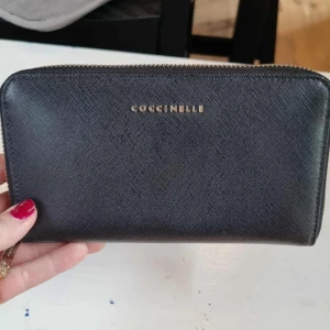 Svart plånbok från Coccinelle i skinn - Stilren svart plånbok från Coccinelle i äkta skinn med guldfärgad dragkedja. Plånboken har flera fack för kort, myntficka med dragkedja och ett fönsterfack för ID. Perfekt för dig som vill ha ordning och snygg design i väskan.