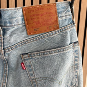 Levi's 501 ljusblå jeans W27 L30 - Klassiska Levi's 501 jeans i ljusblå denim med raka ben och femficksmodell. Byxorna har den ikoniska läderpatchen bak och röda Levi's-tag på bakfickan. Tillverkade i bomull med snygga kopparfärgade nitar och knappgylf. Perfekta för en avslappnad stil.