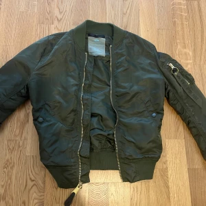 Grön bomberjacka Alpha Industries S - Säljer en klassisk mörkgrön bomberjacka från Alpha Industries i storlek S. Jackan har guldiga dragkedjor, ribbade muddar och den ikoniska 'Remove Before Flight'-taggen på ärmen. Tillverkad i slitstark polyester och har flera fickor för en cool och funktionell look.