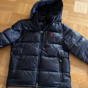 Marinblå pufferjacka Polo Ralph Lauren M - Marinblå pufferjacka från Polo Ralph Lauren med huva och dragkedjor. Jackan har en röd broderad logga på bröstet och flera praktiska fickor med dragkedja. Perfekt för kalla dagar och har en clean, sportig look.