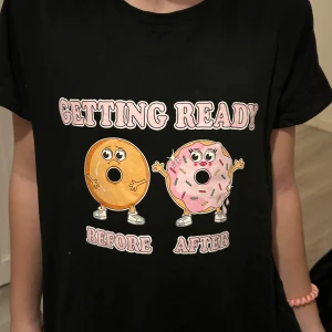 Svart t-shirt med donut-tryck FB Sister - Svart t-shirt från FB Sister i storlek S med ett färgglatt tryck av två donuts och texten 'Getting Ready Before After' på framsidan. T-shirten har korta ärmar och en klassisk passform. Materialet är mjuk bomull som känns skönt mot huden.