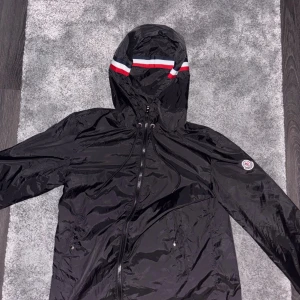 Svart vindjacka från Moncler med huva - Svart vindjacka från Moncler med huva och dragkedja framtill. Jackan har Moncler-logga på ärmen och snygga röd, vit och blå detaljer på huvkanten. Tillverkad i glansig polyester och har snörning vid huvan samt fickor med dragkedja.