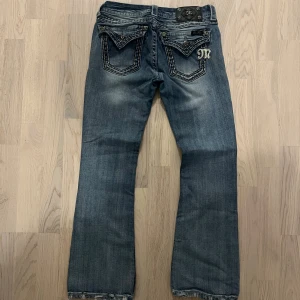 Miss Me blå bootcut jeans med detaljer - Snygga blå jeans från Miss Me med bootcut passform. Jeansen har slitna detaljer, markerade sömmar och dekorativa bakfickor med metallknappar och blingad M-logga. Klassisk femficksmodell i mjuk bomullsdenim. Perfekta för dig som gillar statement-jeans.