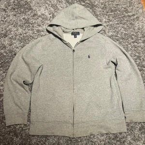 Ralph Lauren Hoodie - Säljer en stilren Ralph Lauren Zipp upp i färgen grå. Storleken är L(14-16) och skicket är som nytt. Passar till både damer och herrar. Vid frågor är det självklart bara att höra av dig😊