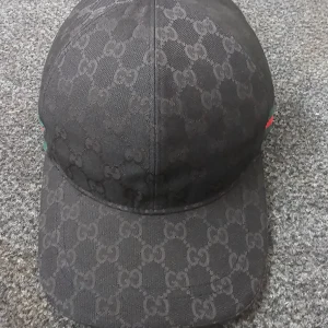 Svart keps från Gucci med GG-mönster - Säljer en svart keps från Gucci med klassiskt GG-monogram över hela kepsen. Kepsen har en böjd skärm, justerbart spänne bak i skinn och en grön-röd rand längs kanten. Tillverkad i bomull och detaljer i skinn. Perfekt för dig som vill ha en lyxig och stilren accessoar.