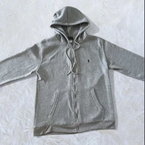 Grå hoodie från Polo Ralph Lauren - Snygg grå hoodie från Polo Ralph Lauren med klassisk broderad logga på bröstet. Tröjan har huva med snörning, dragkedja framtill och två praktiska fickor. Mjuk bomullsmix som är perfekt för chill dagar. Passar dig som gillar stilren och enkel design.