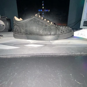 Christian Louboutin svarta sneakers med nitar - Svarta sneakers från Christian Louboutin med ikoniska röda sulor och svarta nitar på tå och häl. Skorna har mönstrad ovandel i skinn och svarta skosnören. Insidan är ljusbeige och märkt med Louboutin Paris.