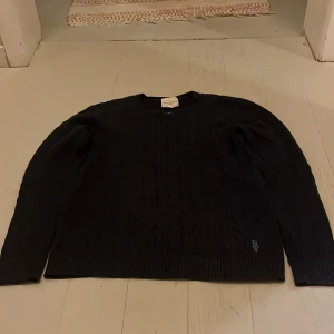 Svart kabelstickad tröja Holly & Whyte - Svart kabelstickad tröja från Holly & Whyte by Lindex med rund hals och ribbade muddar. Tröjan har ett broderat ljusblått logotypmärke vid nederkanten och är långärmad. Perfekt för dig som gillar klassisk stil med en twist.