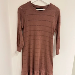 Stickad klänning från Vero Moda - Superfin stickad klänning från Vero Moda i en varm nyans. Modellen har trekvartsärm, rund halsringning och volangkant nertill. Tunt stickat material med diskreta horisontella ränder som ger en snygg struktur. Perfekt för dig som gillar en avslappnad men ändå söt stil.