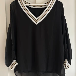 Svart blus med randiga detaljer - Svart blus i luftig polyester med v-ringad hals och vita samt beige randiga muddar vid hals och ärmslut. Blusen har långärmade, lätt transparenta ärmar och loose passform. Perfekt för dig som gillar stilrena och sportiga vibbar.