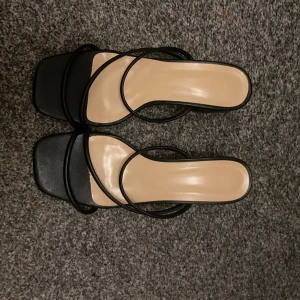 Svarta sandaler med låg klack - Stilrena svarta sandaler med fyrkantig tå och tunna remmar. Skorna har en låg klack och platt sula, vilket ger en modern och minimalistisk look. Perfekta för dig som gillar enkel men trendig design. Materialet är syntet och färgen är klassiskt svart.