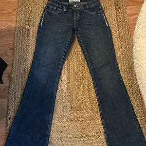 Bootcut jeans med nitar från Bershka - Mörkblå bootcut jeans från Bershka med snygga detaljer i form av nitar och beige broderi på bakfickorna. Klassisk femficksmodell med låg midja och markerade sömmar. Perfekta för dig som vill sticka ut med coola detaljer.