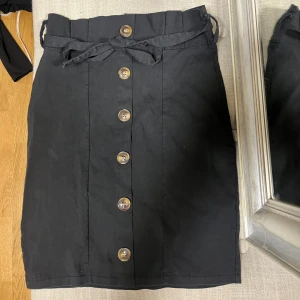 Svart kortkjol med knappar och bälte - Svart kortkjol med snygga knappar framtill och ett matchande bälte i midjan. Kjolen har en rak passform och är tillverkad i ett mjukt bomullsmaterial. Perfekt för dig som gillar stilrena och enkla plagg med en twist. Den andra kjolen har silverfärgade knappar och är i mockaliknande material.