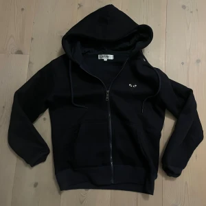 Mörk blå hoodie Comme des Garçons Play S - Svart hoodie från Comme des Garçons Play med dragkedja, huva och snörning. Ikoniskt hjärta med ögon på bröstet och stort hjärta på ryggen. Mjuk bomullskänsla och känguruficka framtill. Perfekt för dig som gillar streetwear och stilrena detaljer.