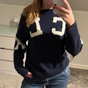 Mörkblå stickad tröja med UCLA-tryck - Snygg mörkblå stickad tröja från UCLA med stora vita bokstäver på bröstet och ärmarna. Tröjan har rund hals, ribbade muddar och en loose passform. Det är en liten storlek men den passar mig som vanligtvis har stl S.