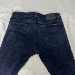 Mörkblå jeans från Nudie Jeans - Snygga mörkblå jeans från Nudie Jeans med klassisk femficksdesign och kontrastsömmar på bakfickorna. Jeansen har en rak passform och är tillverkade i slitstarkt denimtyg. Perfekta för dig som gillar stilrena och tidlösa jeans.