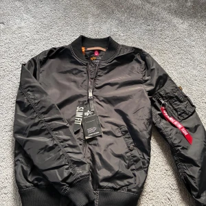Svart bomberjacka Alpha Industries S - Svart slim fit bomberjacka från Alpha Industries med klassisk orange foder, ribbade muddar och dragkedja framtill. På ärmen finns en ficka med röd 'Remove Before Flight'-tag. Jackan har flera fickor och är tillverkad i slitstark polyester. Jacka är helt ny och jag har inte användt den pga för liten, jackan är liten i storleken villket gör att XS passar fint.