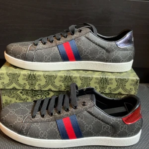 Gucci sneakers med GG-mönster - Snygga sneakers från Gucci i mörkgrått med klassiskt GG-mönster över hela skon. Sidorna har blå och röda ränder, och baksidan har metallicdetaljer i rött och lila med Gucci-logga. Skorna har svarta snören och vit platt sula med präglad undersida.