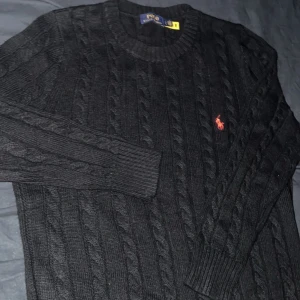 Svart stickad tröja Polo Ralph Lauren - Svart kabelstickad tröja från Polo Ralph Lauren med rund hals och lång ärm. Klassisk röd broderad logga på bröstet. Tröjan har ribbade muddar och är gjord i mjukt stickat material som känns skönt mot huden.