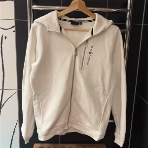 Sail Racing hoodie - Sail racing hoodie använd 1 gång och tvättad jätte bra skick som ny. Storlek 170/XsVit zip-hoodie från Sail Racing. Märkets logga är broderat på vänster bröst och en mindre logotyp längst upp på huvan. Fickor på båda sidor av tröjan samt en ficka med dragkedja bredvid loggan på bröstet. Ribbstickad mudd finns både i ärmslutet och i midjan på denna huvtröja. 