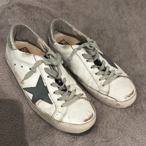 Golden Goose Superstar sneakers vit/silver - Golden Goose Superstar sneakers i vitt skinn med grå snörning, svart stjärndetalj på sidan och silverfärgad häl. Dem är slitna och en sula har låsnat men är enkelt att laga