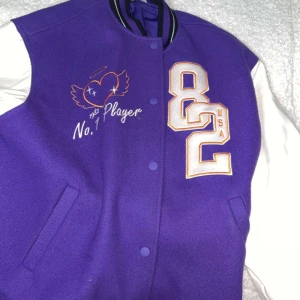 Lila bomberjacka med broderi - Snygg lila bomberjacka från Zoci med vita ärmar och coola broderade detaljer som 'No.1 Player' och stora siffror på bröstet. Jackan har knappar framtill, ribbade muddar och två fickor. Insidan är fodrad i lila och materialet känns som ull och polyester.