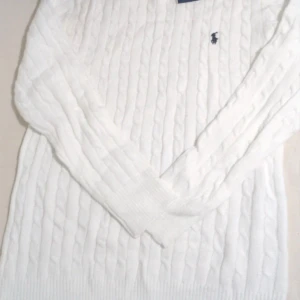 Vit kabelstickad tröja Polo Ralph Lauren - Vit kabelstickad tröja från Polo Ralph Lauren med klassisk broderad logga på bröstet. Tröjan har långärmad design, ribbade muddar och rund hals. Perfekt för dig som gillar stilrena och tidlösa plagg med premiumkänsla.