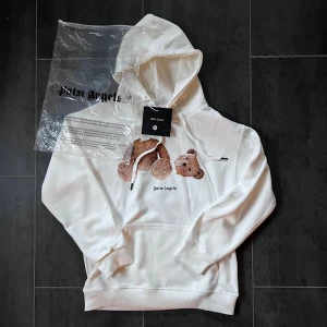 Palm Angels vit hoodie med björn - Palm Angels hoodie i vit färg med stor huva och snörning. Framsidan har ett broderat motiv av en nallebjörn och Palm Angels-logga, medan baksidan har Palm Angels tryckt i svart. Tröjan har ribbade muddar och magficka. Perfekt för dig som gillar streetwear och coola detaljer.
