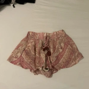 Mönstrade shorts med snörning - Säljer ett par luftiga shorts i ljusrosa och beige med paisleyinspirerat mönster. De har bred resår i midjan och dekorativ snörning med tofsar och pärlor framtill. Materialet är tunt och flowy, perfekt för varma dagar. Plagget har använts ca 2-3 gånger! Pris kan diskuteras 🥰