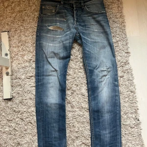 Blå dondup jeans med slitningar - Säljer ett par blå dondup i modellen mius jeans med snygga slitningar och hål på benen. Jeansen har klassisk femficksdesign, raka ben och detaljerade sömmar. Perfekt för dig som gillar en avslappnad och trendig look.