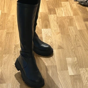 Svarta höga boots i skinn - Säljer ett par svarta höga boots med chunky sula och rund tå. Skorna är i skinn och har en stilren, minimalistisk design utan synliga detaljer. Perfekta för dig som vill ha en edgy och trendig look till höst och vinter.