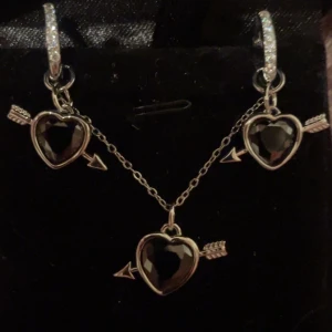 Halsband och örhängen med svarta hjärtan silver - Set med halsband och örhängen i silverfärg med svarta hjärtformade stenar och pil-detaljer. Örhängena har glittrande stenar på ringarna och hjärtan med pil genom. Halsbandet har matchande hänge med samma design. Rostfria