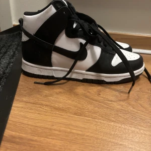 Nike Dunk High svart/vit sneakers - Endast använt 2-3 gånger. Säljer ett par Nike Dunk High sneakers i klassisk svart och vit färg. Skorna har svart swoosh, svarta snören och vit tåbox med perforeringar. Ovandelen är i skinn och sulan är platt och vit med svart yttersula. Perfekt för dig som gillar streetstyle och vill sticka ut.