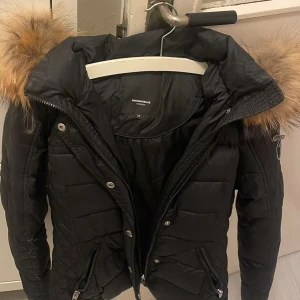 Svart Rockandblue dunjacka med päls - Snygg svart dunjacka från Rockandblue med stor fluffig päls runt huvan. Jackan har quiltade detaljer, dragkedjor på fickorna och silverfärgade knappar. Perfekt för kalla dagar och ger en riktigt lyxig vibe.