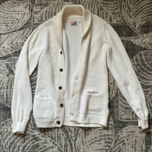 Vit kofta med stickad struktur - Säljer en vit kofta med snygg stickad struktur framtill, ribbad krage och muddar. Koftan har bruna knappar och två fickor framtill. Perfekt för lager-på-lager och chill stil.