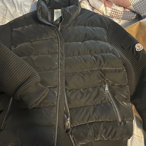 Svart pufferjacka från Moncler - Svart pufferjacka från Moncler med quiltad front och stickade ärmar. Jackan har hög krage, två dragkedjefickor och Moncler-logga på ärmen. Materialet är polyester och stickad textil, perfekt för kalla dagar. Snygg och modern look med detaljer som dragkedja och ribbade muddar.