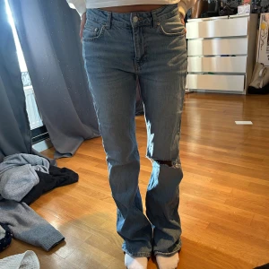 Bootcut jeans med hål på knät - Säljer ett par ljusblå jeans med raka ben och klassisk femficksdesign. Jeansen har hög midja och ett stort hål på ena knät för en cool, avslappnad look. Tillverkade i denim med en skön passform som sitter snyggt på kroppen.