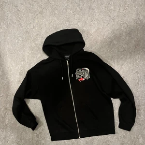 Supply & Demand NYC - Svart hoodie från Supply & Demand New York City i storlek S. Tröjan har huva med snörning, dragkedja framtill och ett coolt tryck med bokstäver på bröstet. Mjuk insida och klassisk passform, perfekt för chill dagar.