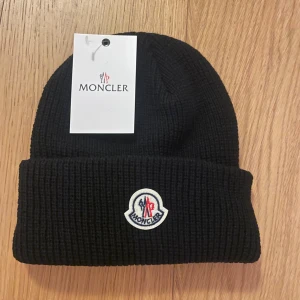 Svart Moncler mössa  - Svart ribbstickad mössa från Moncler med klassisk uppvikt kant och broderad logotyp framtill. Snygg och enkel design som passar perfekt till kalla dagar. Materialet är mjuk ull som håller dig varm och stilen är tidlös.