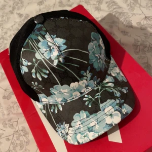  blommig keps med mesh - Snygg keps från  med svart mesh på baksidan och blommigt mönster i blått, vitt och grönt framtill. Justerbar rem bak och klassisk böjd skärm. Tillverkad i bomull och polyester, perfekt för dig som vill sticka ut med designerstil.