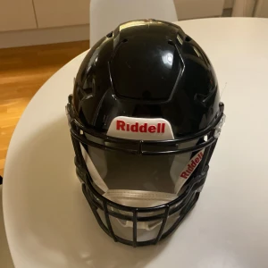 Svart Riddell Legacy hjälm - Säljer en svart amerikansk fotbollshjälm från Riddell Legacy med vit och svart ansiktsgaller, samt tydlig Riddell-logga fram och bak. Hjälmen har vadderad insida och justerbar hakrem för extra komfort och säkerhet. Perfekt för dig som vill ha proffsig look på planen. HELT NY OCH OANVÄND,Pris kan diskuteras
