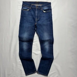Nudie Jeans  - Snygga blå jeans från Nudie Jeans Co med klassisk femficksdesign och orangea kontrastsömmar. Jeansen har en rak passform och subtila slitningar på låren för en snygg look. Hör av dig vid minsta fundering!