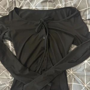 Svart långärmad topp med knyt - Säljer en svart långärmad topp med knyt framtill och knappar längs bröstet. Toppen har en bred, något offshoulder ringning och är gjord i mjukt bomullsmaterial. 