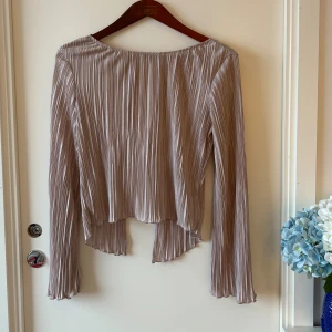 Champagnefärgad omlottblus från NA-KD - Champagne / rosa färgad omlottblus från NA-KD i plisserat, glansigt material. Blusen har långa ärmar, knytning i ryggen och en flowy, asymmetrisk nederkant. Perfekt för dig som gillar stilrena detaljer och en lyxig vibe.