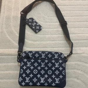 Louis Vuitton svart axelväska monogram - Snygg svart axelväska från Louis Vuitton med klassiskt vitt monogrammönster. Väskan har två stora fack med dragkedja, bred justerbar axelrem och en liten matchande pouch. Tillverkad i skinn med stilren och modern look.
