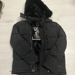 Svart dunjacka Moose Knuckles M - Svart dunjacka från Moose Knuckles med fluffig huva och silvriga metalldetaljer på ärmen. Jackan har dragkedja, flera fickor och mjukt foder. Perfekt för kalla dagar och har en riktigt schysst streetstil. Helt oanvänd, har använt den 2 gånger. 